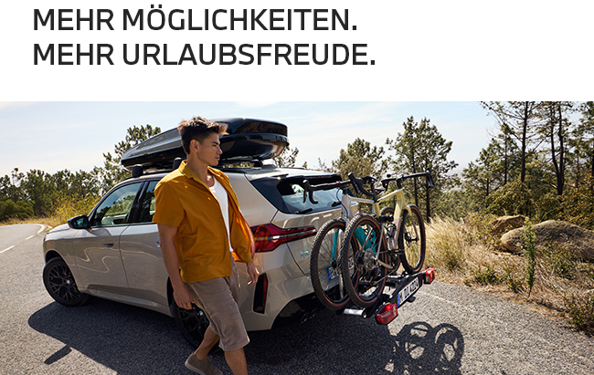 Ein sonniges Werbefoto für Urlaubszubehör: Links steht in dunkler Schrift der Spruch „MEHR MÖGLICHKEITEN. MEHR URLAUBSFREUDE.“  Darunter läuft ein Mann in sommerlicher Kleidung an einem beigefarbenen SUV vorbei, der auf einer Landstraße hält. Das Auto ist voll auf Urlaub ausgerüstet – mit einer schwarzen Dachbox oben drauf und zwei Fahrrädern auf einem Träger am Heck. Im Hintergrund sieht man eine hügelige, mediterrane Landschaft mit Bäumen.