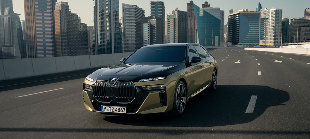 Eine moderne BMW 7er Limousine in markanter Schwarz-Gold-Lackierung fährt auf einer leeren Schnellstraße vor einer beeindruckenden Wolkenkratzer-Skyline.