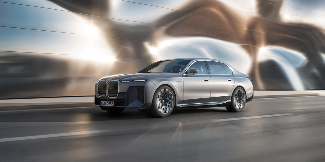 Ein silberner BMW i7 in Fahrt auf einer Straße mit dynamischem Unschärfeeffekt vor einer futuristischen, glänzenden Fassade, die das Licht reflektiert.