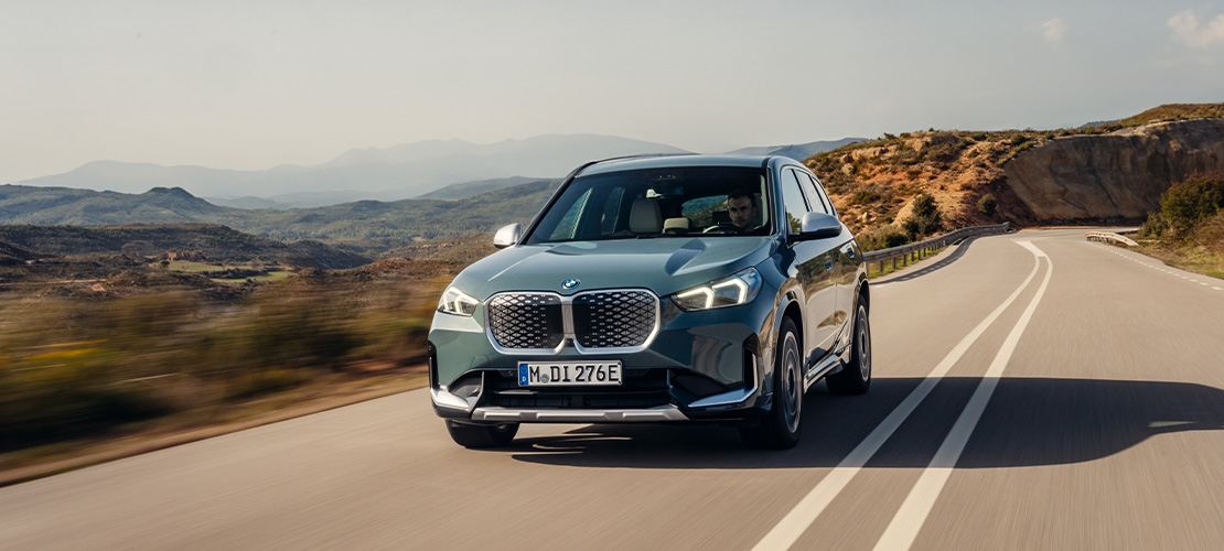 Ein dynamisches Werbefoto zeigt den vollelektrischen BMW iX1 in elegantem Graublau während einer zügigen Fahrt durch eine mediterrane Hügellandschaft. Die markante Front mit der geschlossenen Niere und das Münchner E-Kennzeichen unterstreichen das moderne, emissionsfreie Design, während die leichte Bewegungsunschärfe auf der kurvigen Landstraße Agilität und Fahrfreude vermittelt.