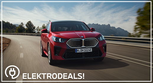 Vorderansicht eines roten BMW iX2 Elektro-Crossovers mit Kennzeichen 'MDH 1024E', der auf einer Landstraße in Richtung Berge fährt. Mit 'Elektrodeals!'-Logo."