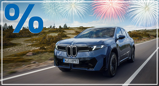 Diese Werbegrafik zeigt einen vollelektrischen BMW iX3 in einem kräftigen Blau, der zügig auf einer asphaltierten Landstraße fährt. Das Fahrzeug ist in einer dynamischen Dreiviertelansicht von vorne abgebildet, während es eine offene, leicht hügelige Landschaft mit niedriger Vegetation unter einem hellen Tageshimmel durchquert.  Der obere Bildrand wird von vier großen, farbenfrohen Feuerwerksexplosionen in Weiß-, Blau- und Rottönen geziert, die sich über die gesamte Breite erstrecken. Das für diese Kampagne charakteristische Designmerkmal – das große blaue Prozentzeichen in der oberen linken Ecke sowie der schmale weiße Rahmen – rundet die Grafik ab.