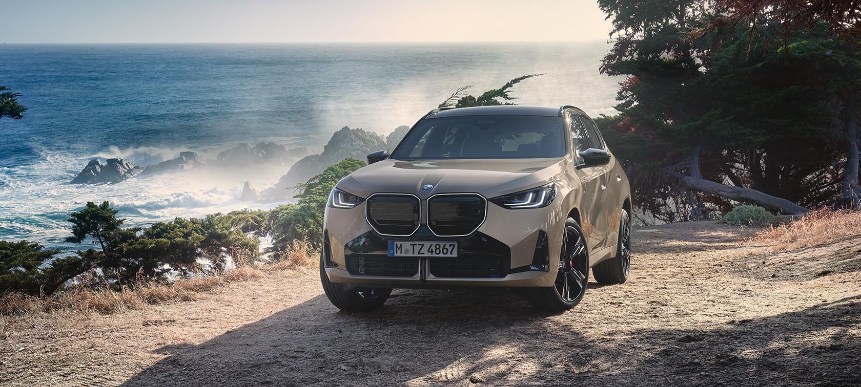 Das Bild zeigt den neuen BMW X3 in einem sandfarbenen Goldton, der auf einer Anhöhe direkt an einer rauen Küste parkt. Im Hintergrund bricht sich das Meer an schroffen Felsen, während das warme Sonnenlicht eine abenteuerliche und zugleich luxuriöse Atmosphäre schafft. Mit seiner markanten, großen Doppelniere und den dunklen Akzenten wirkt das SUV in dieser naturbelassenen Umgebung besonders robust und präsent.
