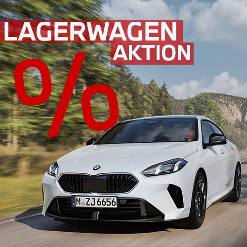 Ein alpinweißes BMW 2er Gran Coupé fährt dynamisch auf einer sonnigen Landstraße vor bewaldeter Bergkulisse. Die sportliche Front mit schwarzen Akzenten steht im Fokus. Das markante rote Banner „Lagerwagen Aktion“ und ein großes Prozentzeichen dominieren die linke obere Bildhälfte und signalisieren ein attraktives Angebot für diesen sportlichen Viertürer.