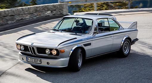 Ein klassischer silberner BMW 3.0 CSL E9 mit Rennstreifen fährt auf einer kurvigen Bergstraße.