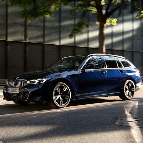  Dunkelblaue BMW 3er Touring Kombi in Bewegung auf einer städtischen Straße, mit einem modernen Gebäude und Bäumen im Hintergrund. Das Bild zeigt das spezifische Modell des Angebots dynamisch