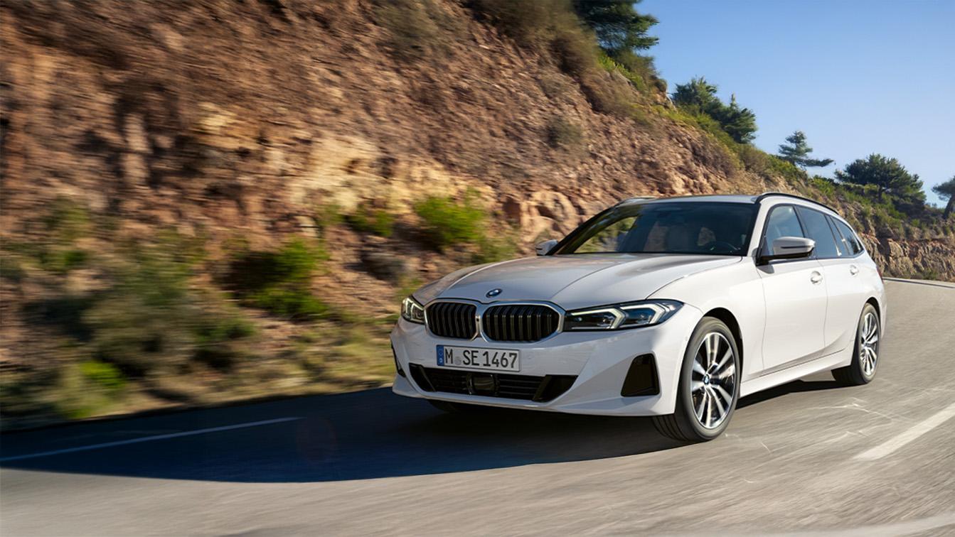 Ein weißer BMW 3er Touring fährt dynamisch durch eine Kurve, wobei die linke Seite des Bildes von einem steilen, felsigen Hang mit grünen Akzenten geprägt ist. Die starke Bewegungsunschärfe des Untergrunds und des Hangs vermittelt ein flüssiges Fahrgefühl bei hoher Geschwindigkeit. Der Wagen ist leicht angewinkelt dargestellt, was die elegante, sportliche Silhouette des Kombis besonders hervorhebt. Das helle Sonnenlicht betont die Konturen der Karosserie und lässt die schmalen LED-Scheinwerfer sowie die dunklen Nierenelemente der Front unter dem strahlend blauen Himmel klar hervortreten.