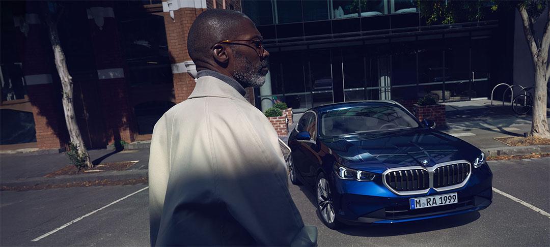 Ein Mann mit Brille und Mantel geht von einem dunkelblauen BMW weg, der vor einem modernen Gebäude parkt.
