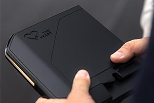 Zwei Hände halten das schwarze, rechteckige Modul mit dem "THE HEART"-Logo. Das Modul ist von der Seite leicht golden oder bronzefarben akzentuiert. Die Person hält es wie ein modernes Tablet oder ein wichtiges technisches Bauteil.
