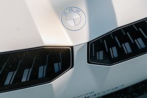 Eine Nahaufnahme der Front eines weißen Fahrzeugs. Im Zentrum ist das BMW-Logo auf der Motorhaube zu sehen. Darunter befinden sich scheinbar die Scheinwerfer oder Lüftungsschlitze, die eine rechteckige Form haben und dunkle, vertikale, beleuchtete oder reflektierende Streifenmuster aufweisen.