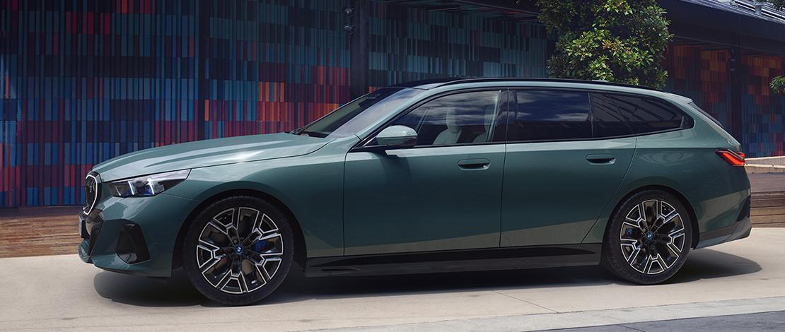  Ein mittelgroßer, dunkelgrüner BMW i5 Touring Kombi ist von der Seite leicht schräg von vorne auf einem hellen Asphaltweg abgebildet. Das Auto hat schwarze Felgen mit blauen Akzenten und getönte Scheiben. Im Hintergrund ist ein modernes Gebäude mit einer farbenfrohen, gemusterten Fassade zu sehen, die aus rechteckigen Elementen in verschiedenen Blau-, Rot- und Grüntönen besteht. Der Himmel ist nicht sichtbar.