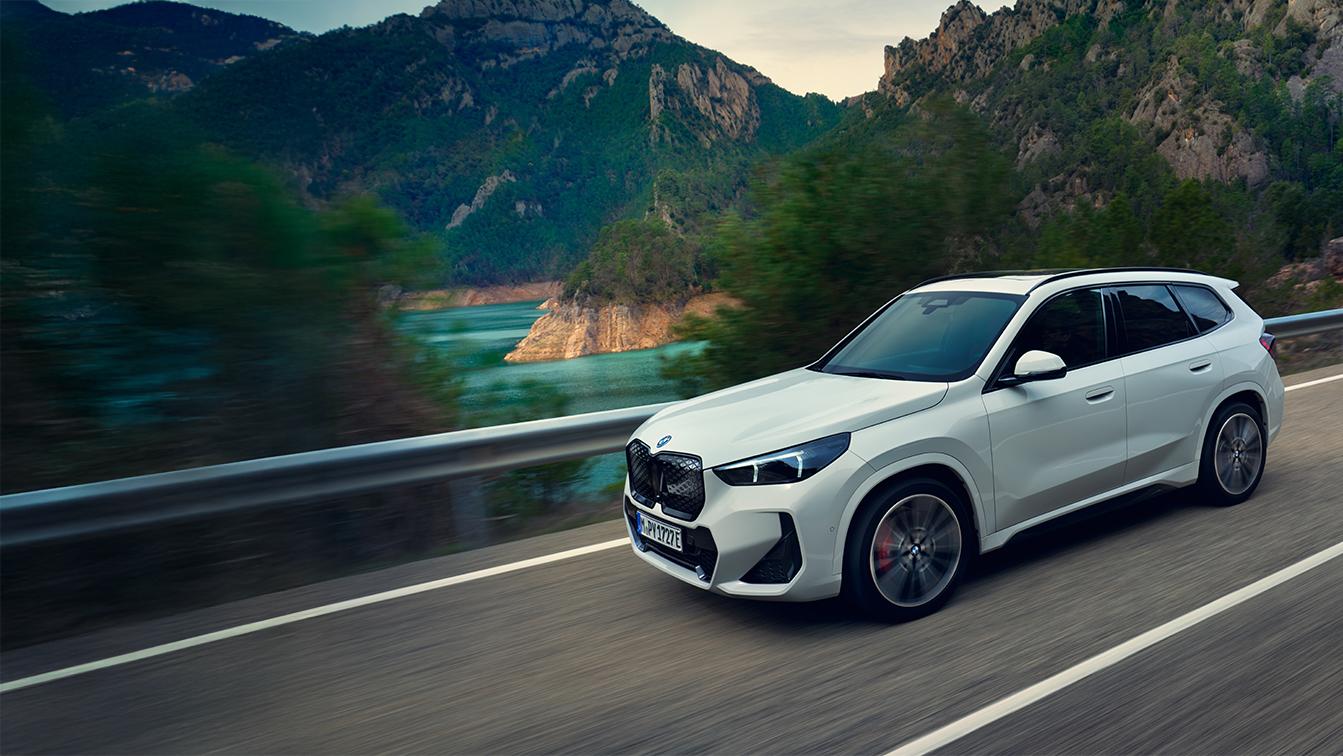 Das Bild BMW iX1 Marketingbonus Q4 2026.jpg zeigt einen weißen BMW iX1 in dynamischer Fahrt auf einer Bergstraße vor einer Kulisse aus Bergen und einem blauen See. Die Bewegungsunschärfe und sportliche Details wie rote Bremssättel betonen das moderne, kraftvolle Design des Elektro-SUVs in der Natur.