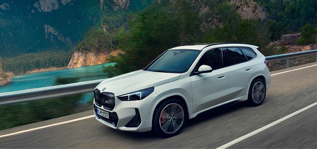 Ein weißer BMW iX1 fährt dynamisch auf einer kurvigen Bergstraße entlang eines tiefblauen Sees. Das SUV ist in einer leichten Schrägansicht von vorne rechts zu sehen, während die unscharfe Umgebung die Geschwindigkeit der Fahrt unterstreicht. Im Hintergrund ragen bewaldete, felsige Berge unter einem klaren Himmel auf, was dem Bild eine abenteuerliche und zugleich elegante Atmosphäre verleiht.