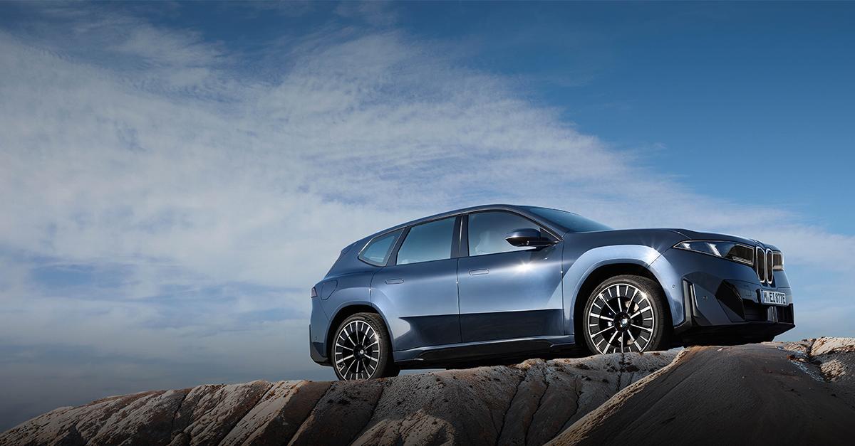 Das Bild zeigt den neuen BMW iX3 in einer imposanten Seitenansicht auf einer felsigen Anhöhe vor einem weiten, hellblauen Himmel. Das vollelektrische SUV besticht durch ein futuristisches Design in einem metallischen Graublau, das durch scharfe Linien, bündige Türgriffe und sehr schmale, markante Scheinwerfer geprägt ist. Die tief stehende Sonne betont die aerodynamische Form und lässt die großen, aufwendig gestalteten Speichenfelgen glänzen. Durch die Aufnahme von einem niedrigen Standpunkt aus wirkt das Fahrzeug besonders kraftvoll und erhaben, fast so, als würde es über der rauen Steinlandschaft thronen.