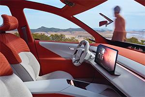 Eine Seitenansicht des Cockpits und des Innenraums mit weiß-orangefarbenen Sitzen. Ein großer Bildschirm ist in der Mitte. Eine Person ist schemenhaft außerhalb des Autos zu sehen.