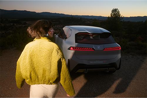 Zeigt das Heck eines hellgrauen Crossover-Fahrzeugs der "Neuen Klasse" bei Sonnenuntergang in einer ländlichen, hügeligen Gegend. Im Vordergrund steht eine Person, die sich vom Betrachter wegdreht und auf das Auto zugeht. Die Person trägt eine voluminöse, hellgelb-grüne Jacke. Die Rücklichter des Autos leuchten rot.