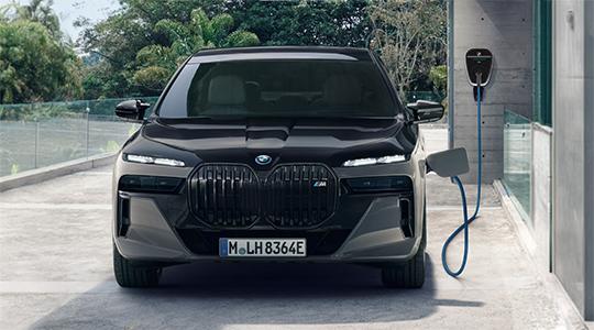 BMW M760 Limousine Das Bild zeigt die Frontansicht einer schwarzen BMW 7er Limousine als Plug-in-Hybrid. Das Auto ist in einer Einfahrt geparkt und wird an einer grauen Wallbox aufgeladen. Die Frontleuchten sind eingeschaltet und die Nieren sind im Vordergrund. Im Hintergrund sind grüne Pflanzen zu sehen.