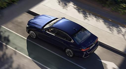 BMW Plug-in-Hybride Das Bild zeigt eine dunkelblaue BMW 5er Limousine Plug-in-Hybrid, die auf einer Straße fährt. Die Aufnahme ist von schräg oben gemacht, sodass das Dach und die Motorhaube gut zu sehen sind. Auf der Straße sind helle Linien und im Hintergrund sind die Schatten von Bäumen zu sehen.