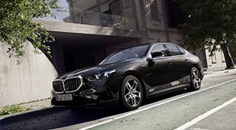 BMW Plug-in-Hybride Das Bild zeigt die Seitenansicht eines dunklen BMW 5er Plug-in-Hybrid, der auf einer Straße geparkt ist. Das Auto steht leicht schräg mit dem vorderen Teil nach links. Im Hintergrund sind ein Gebäude und Bäume zu sehen. Die Frontleuchten des Autos sind eingeschaltet.