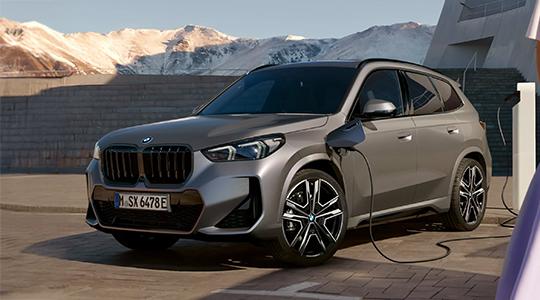 BMW X1 Plug-in-Hybrid Das Bild zeigt die Front eines grauen BMW X1 Plug-in-Hybrid. Das Auto ist draußen auf einem gepflasterten Platz geparkt. Es wird an einer Ladesäule aufgeladen, die sich auf der rechten Seite befindet. Im Hintergrund sind Berge mit Schnee zu sehen.