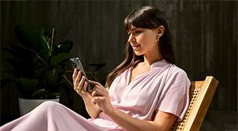 BMW Plug-in-Hybride Das Bild zeigt eine junge Frau in einem hellrosa Kleid, die auf einem Stuhl sitzt. Sie schaut lächelnd auf ihr Smartphone, das sie mit beiden Händen hält. Im Hintergrund ist eine dunkle Wand und eine große grüne Pflanze zu sehen. Die Szene ist von Sonnenlicht beleuchtet.
