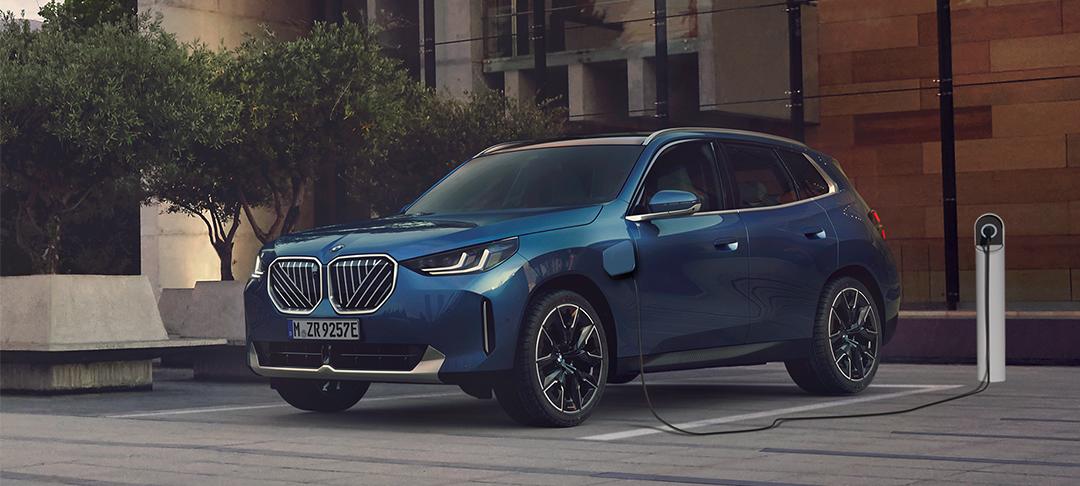 BMW PHEV Fahrzeuge Das Bild zeigt ein Auto, einen BMW X3 Plug-in-Hybrid, das in einem städtischen Umfeld geparkt und an eine Ladesäule angeschlossen ist. Es ist ein blau-metallicfarbenes SUV, das von vorne links zu sehen ist. Die Front des Autos ist geprägt von den markentypischen doppelten Nieren, die hier in einem auffälligen, silbernen Gitterdesign gehalten sind. Links von der Fahrertür ist der Ladeanschluss sichtbar, von dem ein schwarzes Kabel zu einer weißen Ladesäule rechts vom Auto führt. Das Auto steht auf einem gepflasterten Platz neben einem kleinen Baum und einer modernen Sitzbank. Der Hintergrund besteht aus einem Gebäude mit einer strukturierten Fassade. Das Licht ist eher gedämpft, was dem Bild eine ruhige und elegante Atmosphäre verleiht.