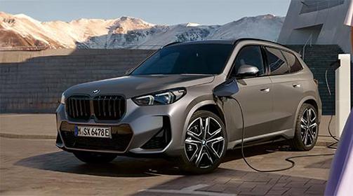 Das Bild zeigt die Front eines grauen BMW X1 Plug-in-Hybrid. Das Auto ist draußen auf einem gepflasterten Platz geparkt. Es wird an einer Ladesäule aufgeladen, die sich auf der rechten Seite befindet. Im Hintergrund sind Berge mit Schnee zu sehen.