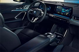 Dies ist eine Innenaufnahme. Man sieht das moderne Armaturenbrett des BMW X1, das große, leicht gebogene digitale Displays für das Cockpit und das Infotainment-System zeigt. Das Lenkrad und Teile der Mittelkonsole mit dem Automatikwählhebel und der Armlehne sind ebenfalls sichtbar. Die Innenausstattung ist dunkel gehalten mit blauen Akzenten.