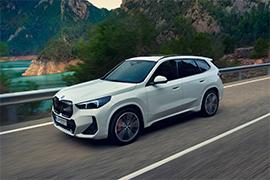 Der gleiche weiße BMW X1 fährt auf einer Landstraße entlang. Im Hintergrund sind grüne, bewaldete Berge und eine Wasserfläche zu sehen, was auf eine Fahrt in landschaftlich reizvoller Umgebung hindeutet. Das Auto ist in Bewegung und von der Seite-vorne aufgenommen.