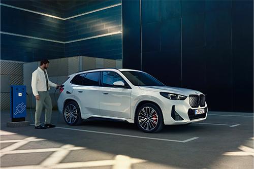 Ein weißer, kompakter SUV (BMW X1) steht auf einem Parkplatz neben einer blauen Ladesäule. Ein Mann in einem hellen Anzug steckt gerade das Ladekabel in die Seite des Autos. Die Szene spielt in einer urbanen Umgebung, möglicherweise in einer Tiefgarage oder vor einem modernen Gebäude mit dunklen und strukturierten Wänden. Der Fokus liegt auf der E-Mobilität.