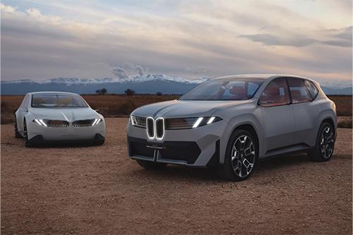 Zwei Fahrzeuge der "Neuen Klasse", eine Limousine und ein Crossover (beide hellgrau), stehen in einer trockenen Landschaft mit Bergen im Hintergrund. Der Crossover ist im Vordergrund.