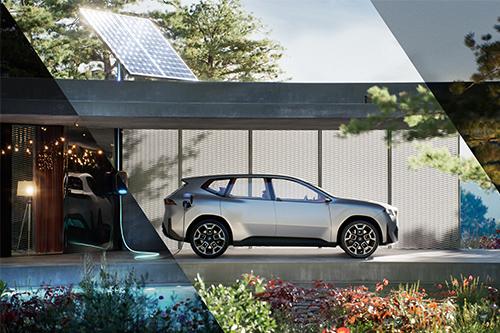 Zeigt einen hellgrauen Crossover der "Neuen Klasse" im Profil, der vor einer modernen Garage oder einem Carport lädt. Im Hintergrund sind Bäume und ein hochgestelltes Solarmodul zu sehen, was auf ein nachhaltiges Energiekonzept hindeutet.