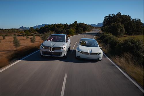 Zeigt zwei Fahrzeuge der "Neuen Klasse" (ein hellgrauer Crossover und eine weiße Limousine) nebeneinander auf einer leeren Landstraße, die sich durch eine sonnige, leicht hügelige Landschaft zieht. Die Autos fahren direkt auf den Betrachter zu.