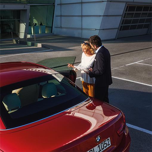 Ein Verkäufer zeigt einer Kundin ein rotes BMW Cabrio vor einem Autohaus.