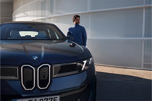 Diese Aufnahme ist eine enge Ansicht der Frontpartie eines BMW Elektroautos, kombiniert mit einer Person. Der Fokus liegt auf der geschlossenen, futuristisch gestalteten Front des dunkelblauen Wagens mit seinen markanten, schmalen LED-Scheinwerfern und der typischen Nierenform. Rechts davon steht ein Mann, der einen dunkelblauen Rollkragenpullover trägt und seitlich in die Ferne blickt. Der Hintergrund besteht aus einer modernen, strukturierten Fassade mit feinen horizontalen Linien, was der Komposition eine urbane Note verleiht.