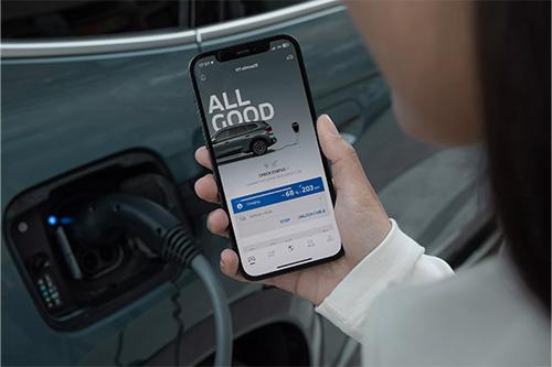 Dies ist eine Nahaufnahme, die die digitale Steuerung des Ladevorgangs illustriert. Im Zentrum hält eine Person ein Smartphone in die Kamera. Auf dem Bildschirm ist eine mobile App von BMW zu sehen, die den Status "ALL GOOD" und Details zum Ladevorgang eines Autos anzeigt. Im unteren Bildbereich sind Optionen wie "CHECK STATUS" und "UNLOCK CABLE" sichtbar. Links unten im Hintergrund ist der beleuchtete Ladeanschluss des Autos zu erkennen, in dem ein dunkles Ladekabel steckt, was signalisiert, dass das Fahrzeug gerade lädt.