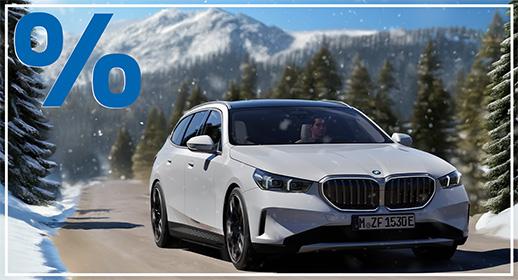 Ein weißer BMW 5er Touring fährt auf einer asphaltierten Straße, die sich durch eine tief verschneite Berglandschaft schlängelt. Im Hintergrund sind hohe, schneebedeckte Gipfel und bewaldete Hänge unter blauem Himmel zu sehen. Das blaue Prozentzeichen (%) füllt die obere linke Ecke und signalisieren einen Rabatt oder eine spezielle Finanzierung.  