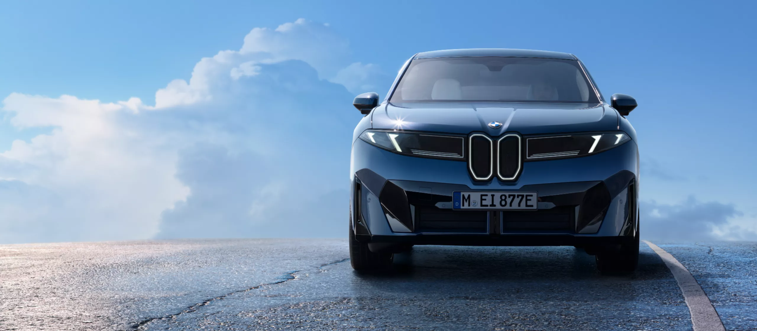 Das Bild zeigt einen blauen BMW iX3 von vorne auf einer nassen, asphaltierten Straße. Der Himmel im Hintergrund ist blau mit weißen Wolken. Das Fahrzeug ist der Hauptfokus des Bildes und die Frontansicht ist gut sichtbar.