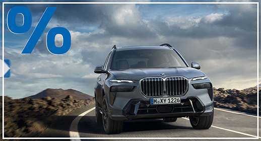Dieses Bild zeigt ein großes, graues SUV, den BMW X7, auf einer kurvigen Straße in einer bergigen Landschaft. Das Auto fährt direkt auf den Betrachter zu. Oben links im Bild ist ein sehr großes, rotes Prozentzeichen (%) platziert. Das Kennzeichen des Autos lautet "M XY 1226".