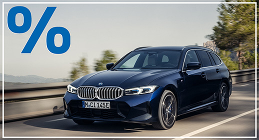  Das Bild zeigt einen dunkelblauen BMW 3er-Reihe Touring (Kombi), der auf einer Landstraße fährt. Das Auto ist schräg von vorne zu sehen. Im Vordergrund ist ein großes, rotes Prozentzeichen (%) platziert. Der Hintergrund zeigt eine bergige Landschaft mit Bäumen unter einem leicht bewölkten Himmel. Das Kennzeichen "M CI 145E" ist auf der vorderen Stoßstange zu sehen.