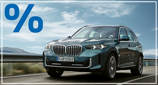 Das Bild zeigt einen dunkelgrünen BMW X5, der auf einer Landstraße fährt. Das Auto ist von vorne zu sehen. Im Vordergrund, über dem Auto, ist ein großes, rotes Prozentzeichen (%) platziert. Der Hintergrund zeigt eine bergige Landschaft unter einem blauen Himmel mit einigen Wolken. Das Kennzeichen "M UP 3185 E" ist auf der vorderen Stoßstange zu sehen.